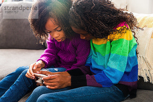 Schwestern in warmer Kleidung mit Smartphone  während sie zu Hause auf dem Sofa sitzen