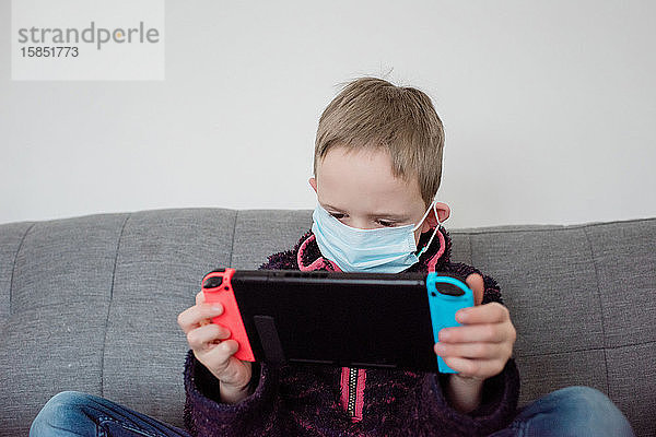 Junge spielt mit einer Spielkonsole mit einer Gesichtsmaske als Schutz