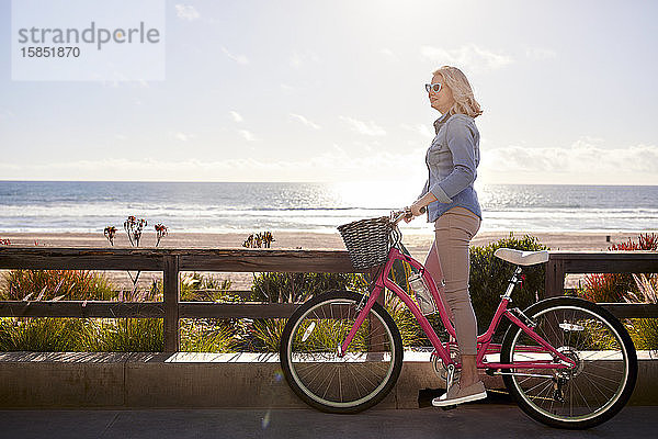 Seitenansicht einer älteren Frau mit Fahrrad  die am Manhattan Beach am sonnigen Tag auf der Straße gegen den Himmel steht