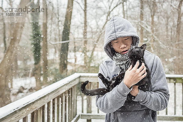 Asiatischer Junge bringt neugierige schwarze Katze an einem verschneiten Tag liebevoll nach draußen