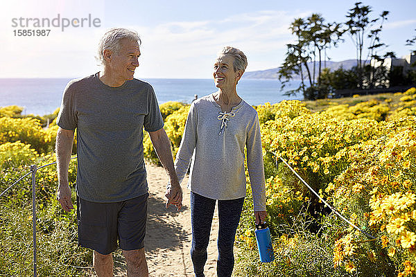Lächelndes älteres Ehepaar hält sich an den Händen  während es am sonnigen Tag auf einem Pfad inmitten von Pflanzen auf einer Klippe am Meer gegen den Himmel geht