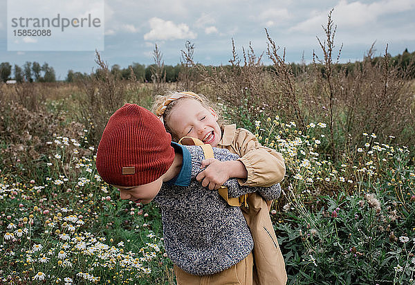 Bruder trägt seine Schwester lachend durch ein Wildblumenfeld