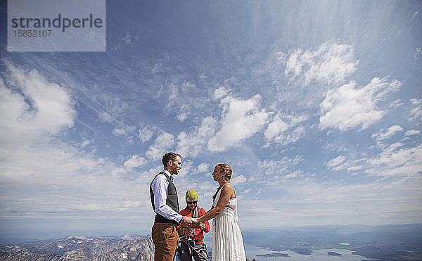 Braut und Bräutigam halten Händchen und heiraten auf einem Berggipfel mit blauem Himmel