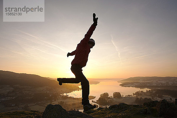 Ein Mann springt vor Freude beim Sonnenaufgang über dem Lake Windermere von Todd Crag über Ambleside  Lake District  Großbritannien.