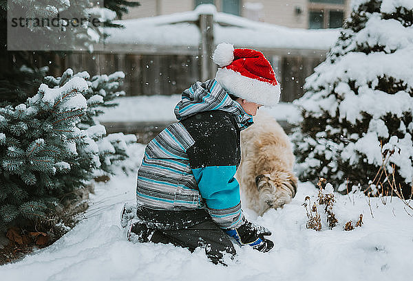 Junge mit Weihnachtsmannmütze  der mit einem Hund im Schnee auf einem Hof spielt.
