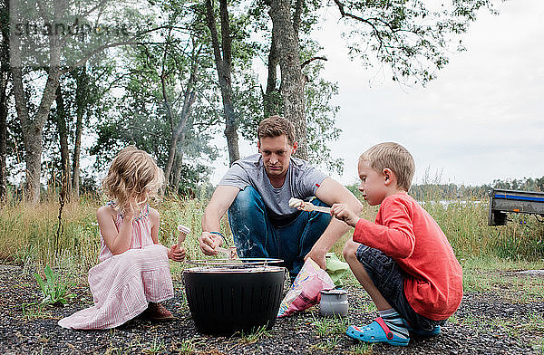 Papa kocht mit seinen Kindern Marshmallows am Lagerfeuer im Freien