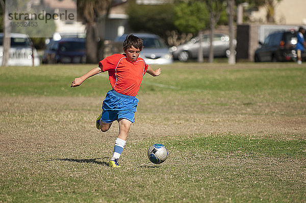 Junger Fussballspieler in rot-blauer Uniform kickt Fussball im Park