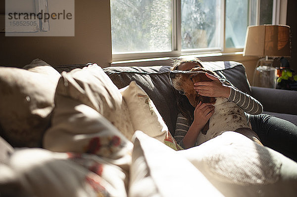 Mädchen umarmt und küsst ihren Hund zu Hause auf dem Sofa in ziemlich hellem Licht