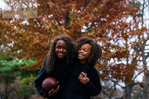 Glückliches Mädchen hält American Football  während sie mit dem Arm um ihre Schwester im Park steht