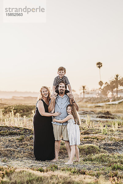 Porträt einer glücklich lächelnden vierköpfigen Familie  die sich bei Sonnenuntergang am Strand umarmt