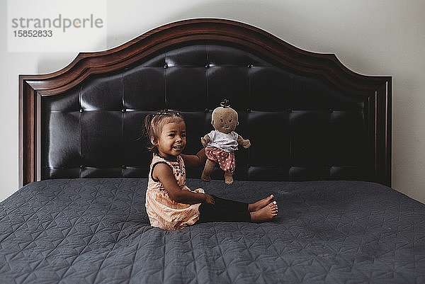 2 Jahre altes gemischtrassiges Mädchen spielt mit Puppe auf dem Bett der Eltern