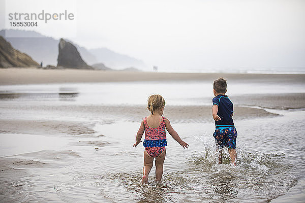 Bruder und Schwester laufen am Strand durch flaches Wasser.