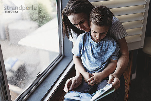 Mutter liest mit ihrem Sohn auf dem Schoß neben dem Fenster