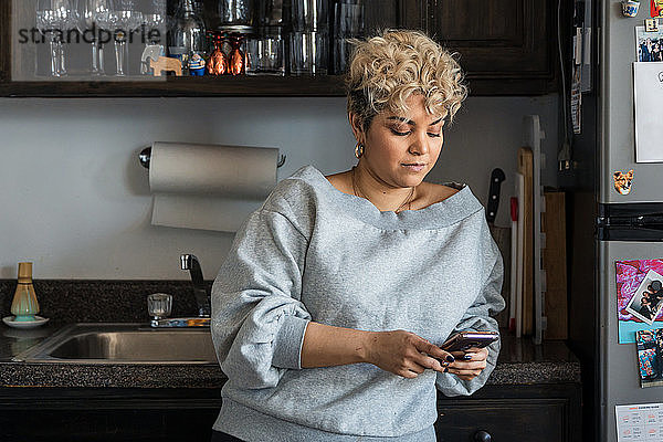 Frau mit kurzen Haaren  die Textnachrichten auf einem Smartphone verschickt  während sie sich zu Hause auf den Küchentisch lehnt