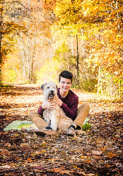 Teenager  der am Herbsttag mit seinem Hund auf einem belaubten Pfad im Wald sitzt.