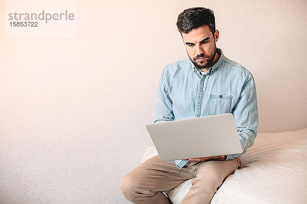 Junger Mann benutzt Laptop-Computer  während er zu Hause an der Wand sitzt