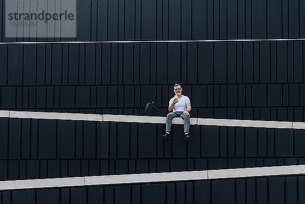 reifer bärtiger Geschäftsmann mit Smartphone  der mit Blick auf die Kamera an der Wand sitzt