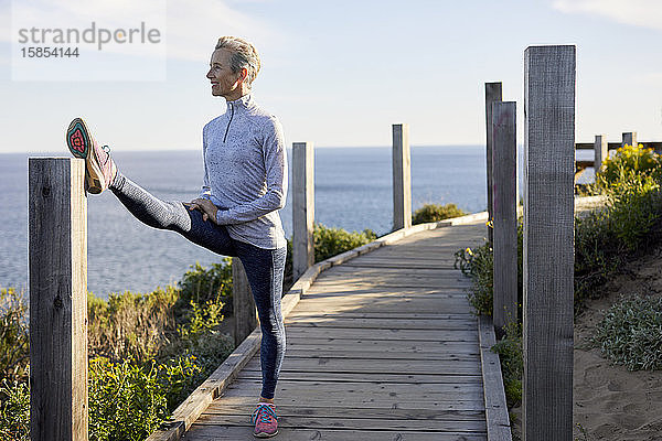 Ältere Frau in voller Länge beim Stretching  während sie auf der Strandpromenade am Meer gegen den Himmel steht