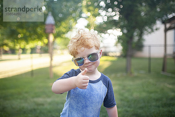 Junge Junge gibt mit Sonnenbrille im Hinterhof stehend einen Daumen hoch