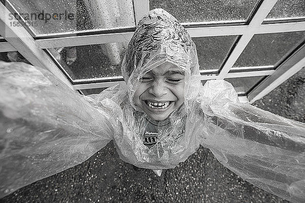 Lächelndes Kind mit Regenmantel im Regen in der Nähe einer Glastür