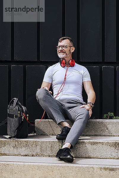 Bärtiger  reifer Mann sitzt mit Kopfhörern auf Stufen und entspannt sich an einem sonnigen Tag
