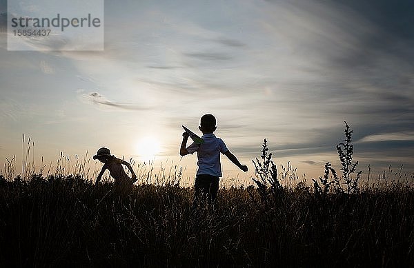 Silhouette von Geschwistern  die bei Sonnenuntergang draußen mit Papierflugzeugen spielen