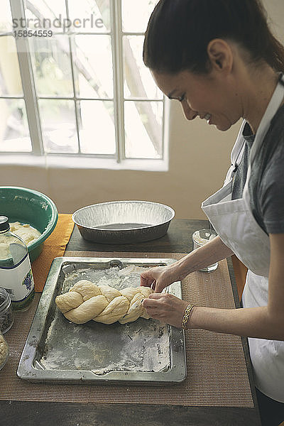Lächelnde junge Frau bereitet zu Hause Challah-Brot aus Teig in einem Tablett auf dem Tisch zu
