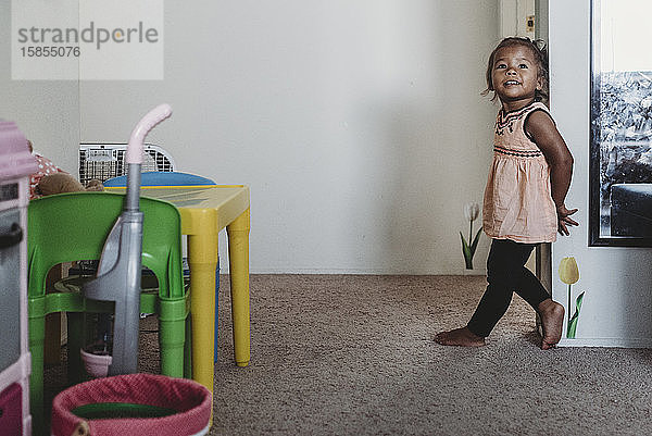 Lächelndes 2 Jahre altes mehrrassiges barfüßiges Mädchen auf Teppich an der Wand stehend