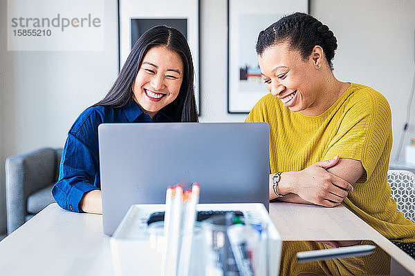 Lächelnde Mitarbeiterinnen diskutieren über Laptop am Tisch im Büro