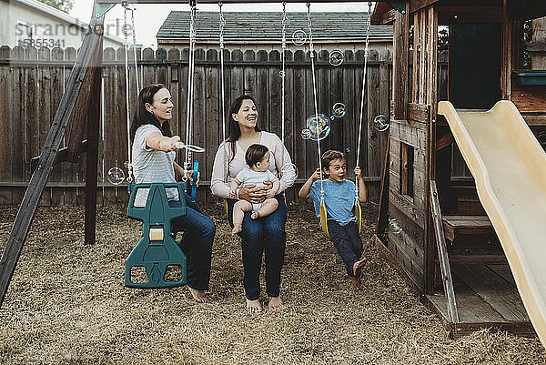 Glückliche Familie mit zwei Müttern auf Schaukeln  die mit Seifenblasen spielen