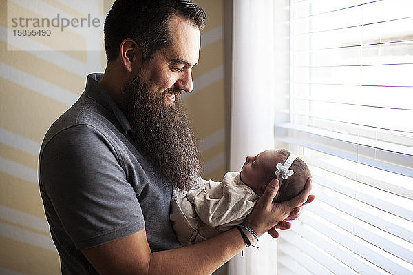 Vater lächelt  während er seine neugeborene Tochter zu Hause am Fenster hält