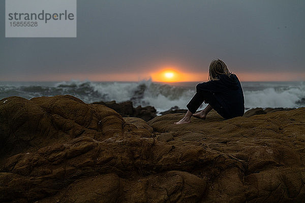 Teenager sitzt an felsiger Küste und schaut bei Sonnenuntergang auf den Ozean