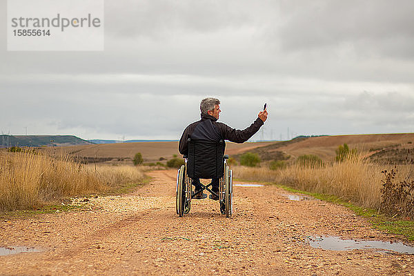 Rückansicht eines behinderten Mannes im Rollstuhl auf einem Weg  der ein Selfie
