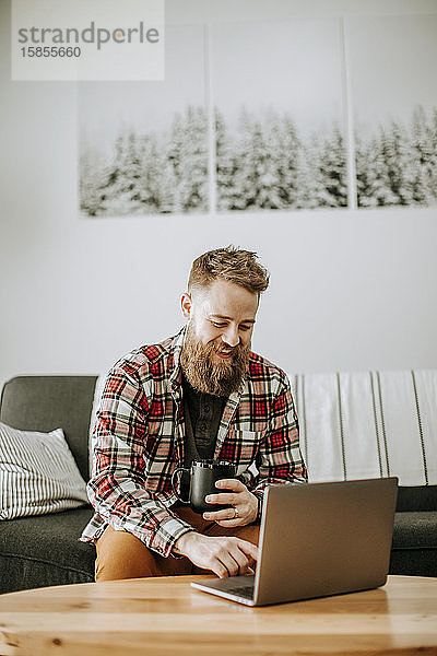 Mann mit Bart hält Tasse Kaffee  während er am Laptop arbeitet