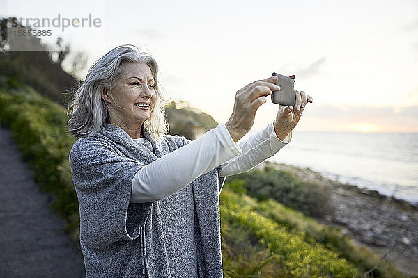 Seitenansicht einer fröhlichen älteren Frau  die mit einem Smartphone am Manhattan Beach bei Sonnenuntergang fotografiert
