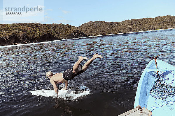 Mann ohne Hemd mit Surfbrett  der bei Sonnenschein gegen den Himmel ins Meer springt