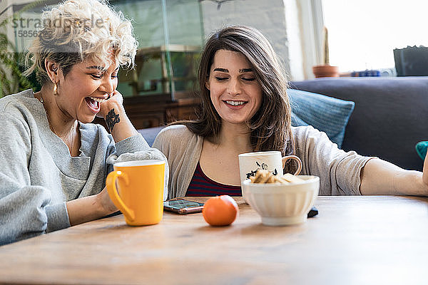 Glückliche weibliche Mitbewohnerinnen benutzen ein Smartphone  während sie Kaffee bei Tisch trinken