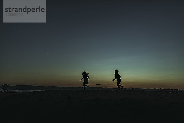 Sihouette von Bruder und Schwester rennen in der Abenddämmerung auf den Ozean zu