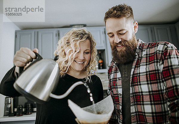 Ein lächelndes junges Paar lässt sich in seiner Küche Kaffee übergießen