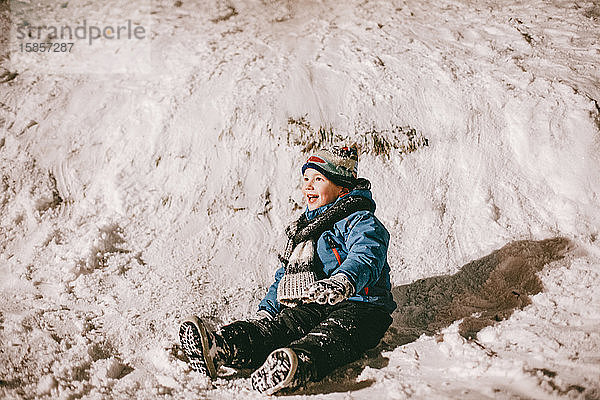 Fröhlicher Junge sitzt im Winter auf Schnee neben einem Hügel