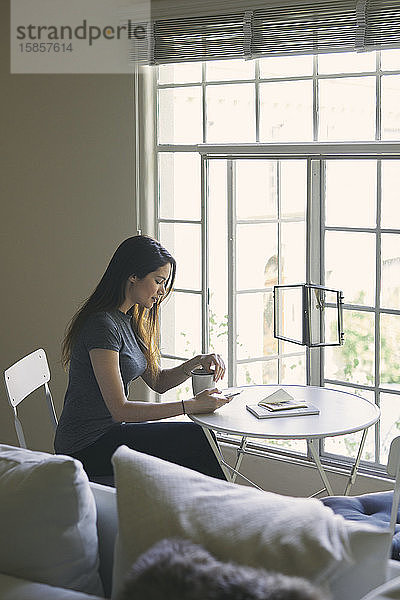Frau benutzt Mobiltelefon  während sie im Wohnzimmer Kaffee am Tisch trinkt