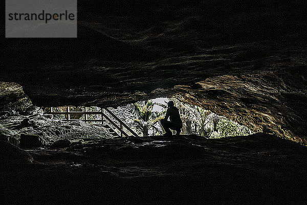 Eine Frau in Silhouette kniet vor dem Eingang einer Höhle in Neuseeland