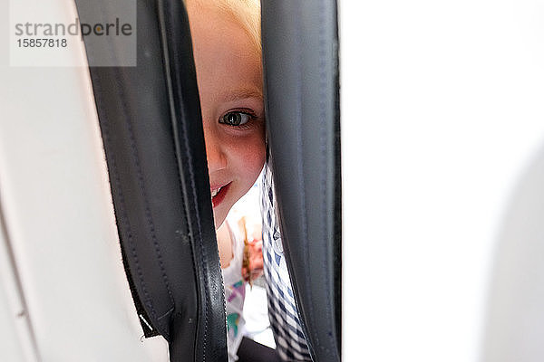 kleines Mädchen  das zwischen den Sitzen im Flugzeug lächelt und in die Kamera schaut