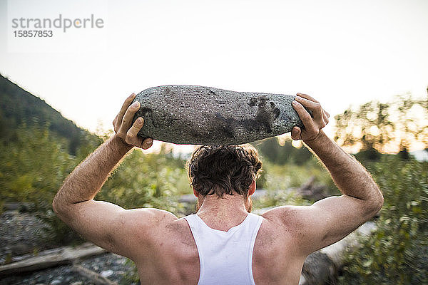 Rückansicht eines Mannes  der während eines Trainings im Freien Felsen über den Kopf hebt.