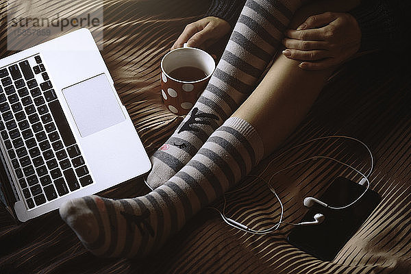 Junges schönes Mädchen in wisitting im Bett mit Tasse Kaffee  Chillen  beobachtend auf dem Bildschirm des Laptops. Winter  gemütlich  Kleidung und Lifestyle-Konzept.