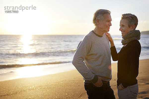 Lächelndes älteres Paar steht sich bei Sonnenuntergang am Strand gegenüber