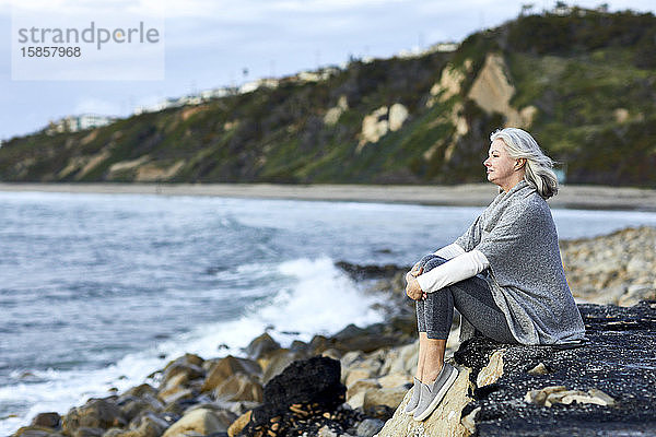 Seitenansicht einer älteren Frau  die auf das Meer schaut  während sie bei Sonnenuntergang auf einem Felsen sitzt