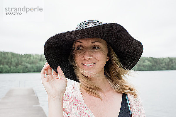 Frau steht an einem windigen Strand und lächelt mit einem Hut auf