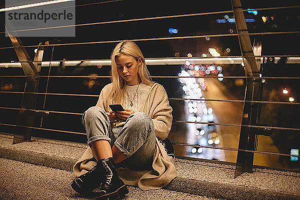 Teenager-Mädchen benutzt Smartphone und sitzt nachts auf einer Brücke in der Stadt