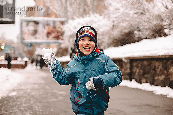 Glücklicher Junge hält Schneeball beim Laufen auf der Straße im Winter